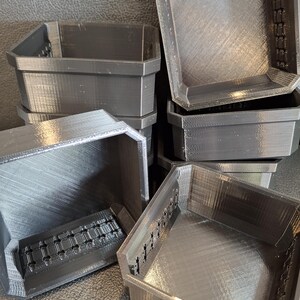 May include: Several dark gray, 3D-printed, octagonal-shaped containers are stacked and arranged. The containers have a textured surface and a series of small holes along the sides. The containers appear to be empty.
