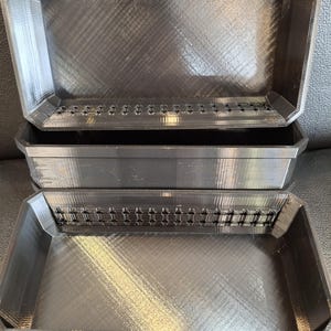 May include: Three black rectangular trays, possibly for storage or organisation. The trays have a textured surface and a series of small holes along the top edge. The trays are stacked on top of each other.