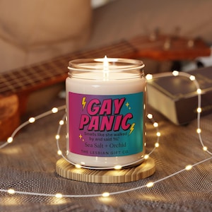 May include: A lit candle in a glass jar with a colorful label that reads "GAY PANIC." The label also states the scent is "Sea Salt + Orchid." The candle sits on a wooden coaster, with string lights and a guitar in the background.