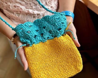 May include: A handmade crochet bag with a bright yellow body and a turquoise blue scalloped top. The bag has a matching turquoise blue strap. The bag is held by a person.