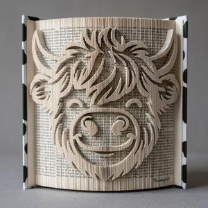 Op de afbeelding: Een gevouwen boekkunstsculptuur met een gedetailleerd gezicht van een Schotse hooglander, gemaakt door de pagina's van een boek nauwkeurig te vouwen. De boekomslag heeft een zwart-wit koeienvlekkenpatroon en de pagina's tonen zichtbare tekst.