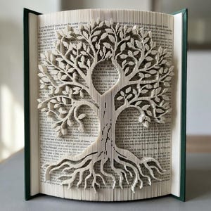 Peut inclure: Sculpture de livre artisanal avec un motif d'arbre découpé dans les pages. Les racines, le tronc, les branches et les feuilles de l'arbre sont finement découpés, créant un effet tridimensionnel. Le livre a une couverture verte et est ouvert, affichant l'œuvre d'art détaillée.