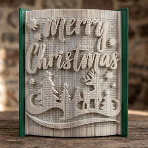 May include: A folded book art sculpture featuring the text Merry Christmas in a cursive font, accompanied by folded paper silhouettes of pine trees, a reindeer, and small stars. The book has dark green covers and cream-coloured pages.
