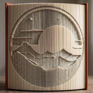 May include: A book sculpture featuring folded pages that create a circular relief of a landscape with a large sun rising over mountain peaks. The book has a rust-coloured spine and the paper edges are precisely manipulated to form the scene.