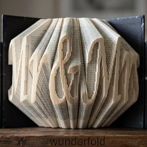 May include: A folded book art sculpture displaying the text Mr & Mrs, created by precisely folding individual pages of a book. The sculpture is positioned between two black marble bookends, showcasing the intricate paper edges and text.