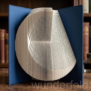 Op de afbeelding: Een boeksculptuur met pagina's die zijn gevouwen tot een cirkelvorm, geplaatst in een donkerblauwe harde kaft. De binnenpagina's zijn dicht op elkaar geplaatst en verticaal uitgelijnd, wat een geometrisch patroon vormt dat lijkt op een halve maan.
