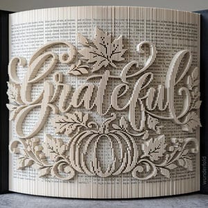 以下が含まれることがあります： ページを折り込んで「Grateful」という文字を形作ったブックアート。中央にはカボチャ、周囲にはカエデの葉がデザインされています。正確なページの折り込みにより、開いた本の上に立体的な効果が生まれています。