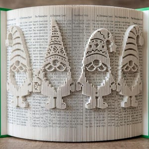 May include: A folded book art sculpture featuring four gnomes standing in a row. The pages are precisely folded to create the silhouettes of the gnomes, each wearing a pointed hat. The book has bright green covers visible on the left and right sides.