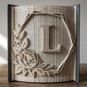 Op de afbeelding: Een gevouwen boekkunstsculptuur met de letter L gecentreerd in een geometrisch kader. Het ontwerp bevat een gedetailleerd bloemstuk met bladeren en een bloeiende bloem aan de linkerkant, gecreëerd door de pagina's van het boek nauwkeurig te vouwen.