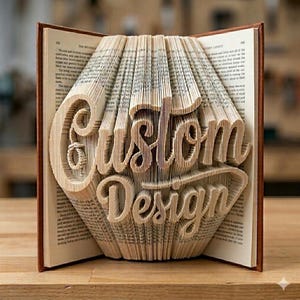 Könnte beinhalten: Eine Buchskulptur mit gefalteten Seiten, die die Wörter Custom Design in Schreibschrift bilden. Das Buch hat einen braunen Einband und weiße Papierseiten mit schwarzem Drucktext, der auf den ungefalteten Abschnitten des offenen Buches sichtbar ist.