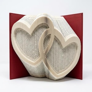 May include: A book sculpture featuring folded pages that form two interlocking heart shapes. The book has a deep red hard cover that is open, revealing the cream-colored paper pages which are precisely creased to create the heart design.