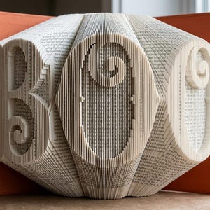 May include: A folded book art sculpture featuring the word BOO created by precisely folding the pages of a book. The sculpture has an orange hardback cover visible behind the white paper pages, which are arranged to form three large letters.