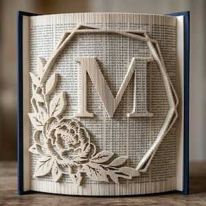 May include: A folded book art sculpture featuring the letter M centred within a geometric hexagonal frame. The design is accented by a detailed floral arrangement with leaves and a blooming flower, all created by precisely folding the book pages.