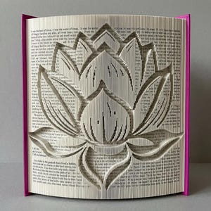Peut inclure: Une sculpture de livre avec un design de pages pliées formant la silhouette d'une fleur de lotus. Les pages sont blanches avec du texte imprimé en noir, et le livre présente des couvertures rose vif visibles sur les côtés gauche et droit.