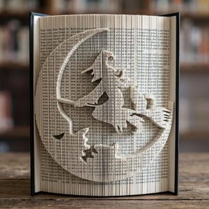 May include: A folded book art sculpture featuring a silhouette of a witch flying on a broomstick in front of a large crescent moon. The pages are precisely folded to create the image, with black covers visible on the left and right sides of the book.