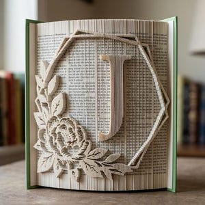 May include: A folded book art sculpture featuring the letter J centred within a geometric frame. The design is accented by a detailed floral arrangement with leaves and petals on the left side, created by precisely folding the pages of the book.