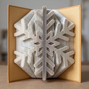 May include: A book sculpture featuring pages folded to create a three-dimensional snowflake design. The book has tan covers and white paper pages with visible black printed text, forming a symmetrical, intricate geometric pattern.