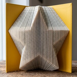 Puede incluir: Una escultura de libro con páginas dobladas para crear una estrella de cinco puntas. El libro está abierto con cubiertas de color amarillo brillante a ambos lados, enmarcando los intrincados pliegues de papel blanco del diseño central.