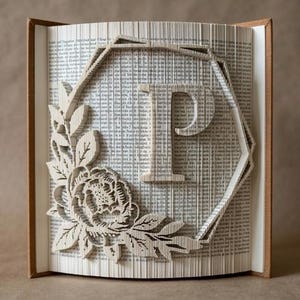 May include: A folded book art sculpture featuring the letter P centered within a geometric hexagonal frame. The design includes a detailed floral arrangement with leaves and a blooming flower on the left side, created by precisely folding pages.