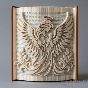 May include: A book sculpture featuring folded pages that form the silhouette of an angel with large wings, a halo, and hands held in a prayer position. The sculpture is created from the pages of a printed book with visible text and brown covers.