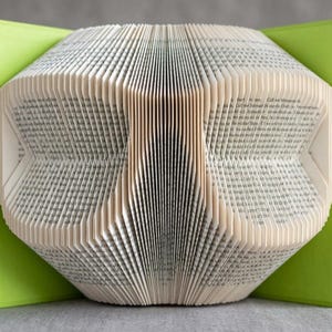 May include: A book sculpture featuring pages folded to create a geometric, symmetrical pattern. The book is held open by bright lime green covers, revealing the printed text on the edges of the white paper pages, forming a structured, three-dimensional shape.