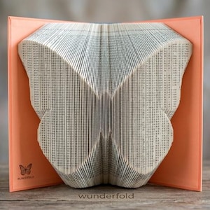 Op de afbeelding: Een boeksculptuur met pagina's die zijn gevouwen in de vorm van een vlinder. Het boek staat tegen een lichtoranje kaft met een klein vlindericoon en de tekst "wunderfold" gedrukt in de linkerbenedenhoek en in het midden onderaan.