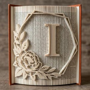 May include: A book art sculpture featuring the letter I folded into the pages. The design includes a geometric hexagonal frame surrounding the letter, accented by a detailed floral arrangement with leaves and a blooming flower on the left side.