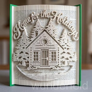 Könnte beinhalten: Eine gefaltete Buchkunst-Skulptur mit dem Text Home for the Holidays. Die Seiten sind kunstvoll gefaltet, um eine dreidimensionale Szene einer Hütte umgeben von Tannenbäumen zu bilden, mit einem grünen Buchrücken an den Seiten.