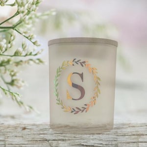 以下が含まれることがあります： つや消しガラスのキャンドルホルダー。装飾的なリースと文字「S」が描かれています。 リースは緑と金の葉で構成されています。 キャンドルホルダーは薄茶色の縁があり、木製の表面に置かれています。