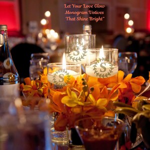 May include: Three lit votive candles in glass cylinders with the letter "S" and a wreath design. The candles are surrounded by orange flowers and are on a table setting. Text above the candles reads "Let Your Love Glow Monogram Votives That Shine Bright."