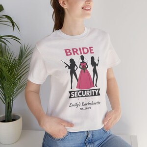 以下が含まれることがあります： ピンクと黒で「BRIDE SECURITY」の文字が入った白いTシャツ。デザインは、ピンクのドレスを着て花を持っている女性と、武器を持っている他の2人の女性のシルエットが特徴です。「Emily's Bachelorette est. 2025」の文字もプリントされています。