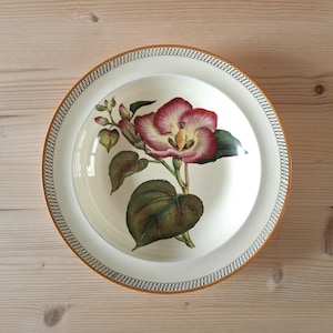 May include: A white ceramic plate with a floral design. The plate features a large, detailed illustration of a flower with red and yellow petals, green leaves, and a brown stem. The rim has a decorative border with a gold and black pattern.