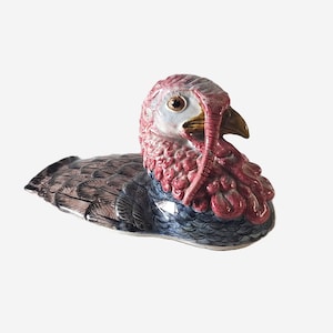 May include: A ceramic turkey ornament with a detailed design. The turkey has a red head and wattle, a brown beak, and a body with blue, brown, and black feathers. The ornament is likely a decorative item.