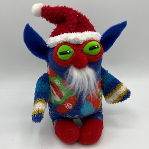 May include: A blue and red Christmas elf plush toy with a white beard and a Santa hat. The elf has green eyes and a red nose.