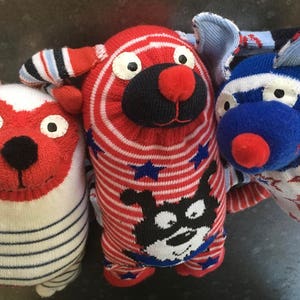 May include: Three handmade sock puppets in red, white, and blue. The puppets are shaped like dogs and have different patterns and designs.