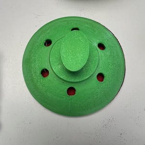 May include: A circular, bright green 3D-printed sanding block featuring a central oval handle and six evenly spaced circular holes around the perimeter. The underside reveals a dark red abrasive surface visible through the ventilation holes.