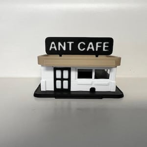 以下が含まれることがあります： 白い建物、ベージュの屋根、黒いベースが特徴の、カフェのミニチュアモデル。上部の看板には白字で「ANT CAFE」と書かれています。カフェには黒いドアと窓があります。