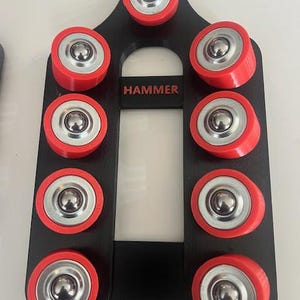 Può includere: Uno strumento nero con la scritta "HAMMER" in rosso. Ha dieci cuscinetti a rulli rossi e argentati attaccati. Lo strumento è progettato per tenere i cuscinetti in posizione.