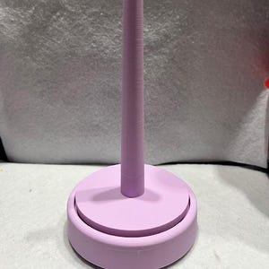 Puede incluir: Un portabobinas de color lavanda con una base redonda y un poste central cilíndrico y alto. La base tiene un borde elevado y el poste está texturizado. El soporte está diseñado para sujetar bobinas de hilo o lana.