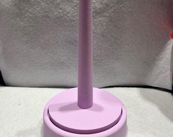 May include: A lilac-coloured spool holder with a round base and a tall, cylindrical centre post. The base has a raised edge, and the post is textured. The holder is designed to hold spools of thread or yarn.