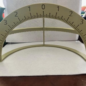 Puede incluir: Un transportador semicircular beige con una escala de 0 a 6 en cada lado. El transportador tiene una parte superior curva y una base plana, con un soporte vertical en el centro. Los números y las marcas están impresos en negro.
