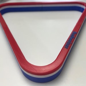 Puede incluir: Un triángulo rojo, blanco y azul para bolas de billar. El soporte tiene un exterior rojo, una banda central blanca y un interior azul. La palabra "Budweiser" está impresa en el lateral del soporte.