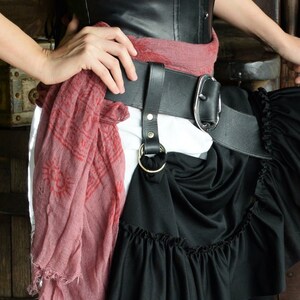 May include: A black leather corset with a red scarf tied around the waist. A black leather belt with a silver buckle is worn over the scarf. A black skirt with ruffles is worn over the belt.