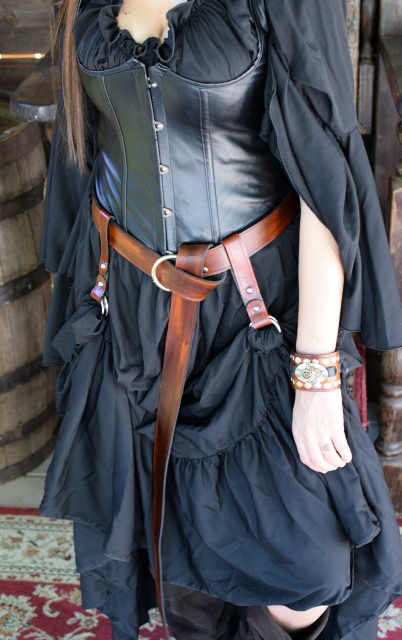 May include: A black leather corset with a brown leather belt and a silver buckle. The corset has a lace trim and is worn over a black skirt.