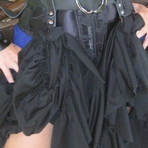 May include: A black corset with a black leather belt and a silver buckle. The corset is worn over a black skirt with ruffles.