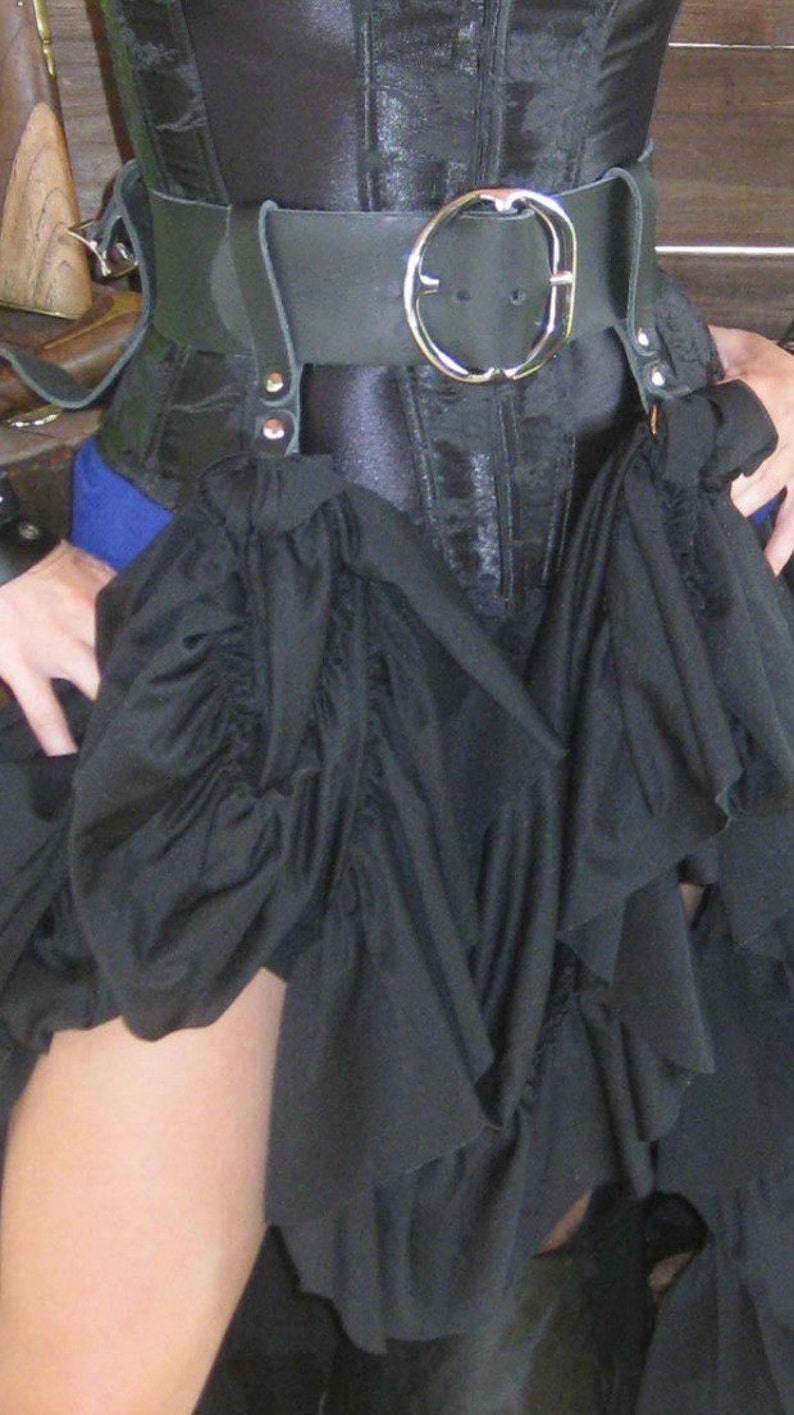 May include: A black corset with a black leather belt and a silver buckle. The corset is worn over a black skirt with ruffles.