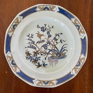 Peut inclure: Assiette en céramique blanche avec une bordure géométrique bleue et dorée. Le centre présente une illustration détaillée d'un oiseau, de fleurs et de feuillage dans des tons de bleu, marron et or. L'assiette a un bord festonné.