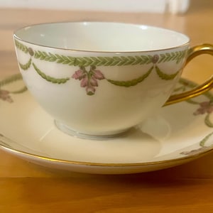 Puede incluir: Un juego de taza y platillo blancos con ribetes dorados. La taza tiene un asa dorada y está decorada con guirnaldas florales verdes y rosas. El platillo refleja el diseño de la taza, creando una estética refinada. Un juego de té clásico.