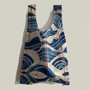 Puede incluir: Bolsa de compras reutilizable con un patrón de olas azul y blanco y rayas verticales rojas. La bolsa tiene dos asas y está hecha de un material ligero. El diseño recuerda al arte japonés.