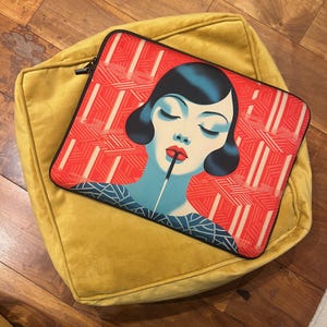 以下が含まれることがあります： 黒いジッパーが付いたマスタードイエローのビーンバッグチェア。 閉じた目と赤い口紅をした女性のレトロなイラストが特徴の赤と白のラップトップスリーブが、ビーンバッグの上に置かれています。